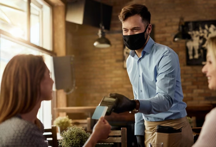 The man wearing a face mask collects the payment from the consumer during the hospitality sales training session.