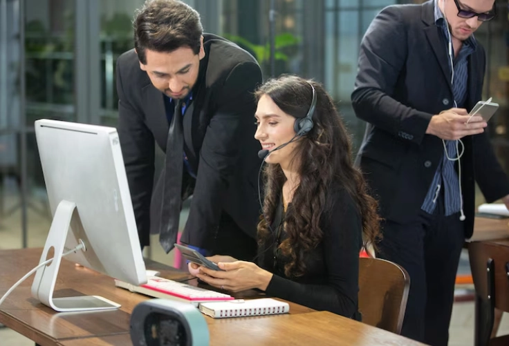 Three people communicate with each other while seated at a desk using computers in an office setting