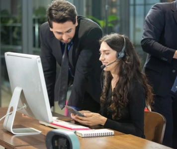 Three people communicate with each other while seated at a desk using computers in an office setting